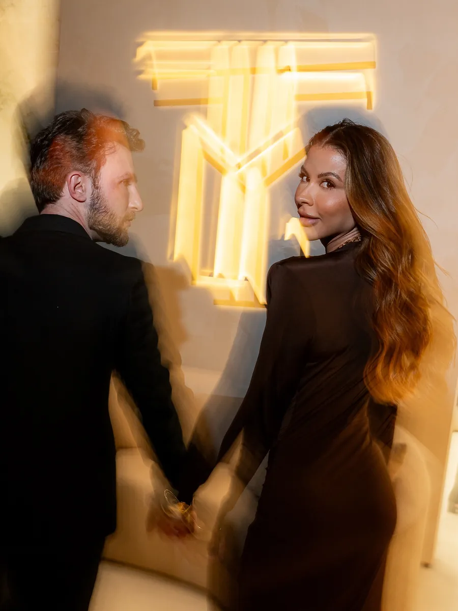 Tommaso e Amanda de mãos dadas diante do monograma dourado, na noite de inauguração